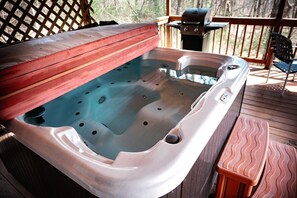 A hot tub on a wooden deck with a grill and chairs in the background. - Lazy Lane Cabins - Cardinal's Crest (Logan)