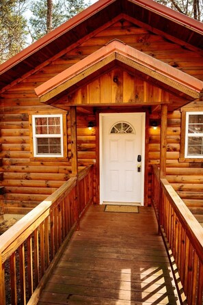 Una cabaña de troncos con una puerta central, una pequeña ventana a cada lado y una terraza de madera que conduce a la entrada.