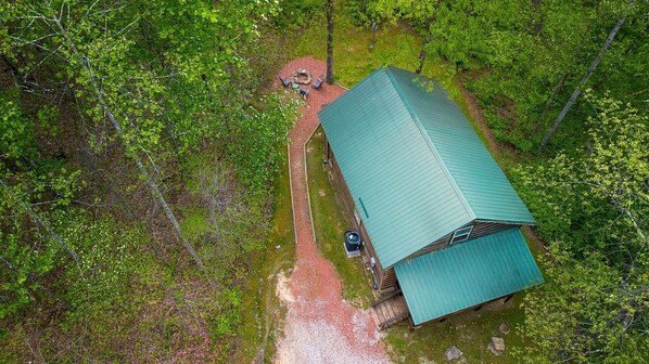 An aerial view of a cabin with a green roof, surrounded by dense forest.