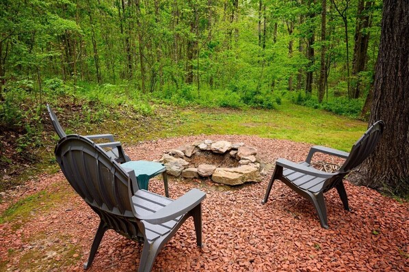A forested area with a fire pit and two Adirondack chairs.