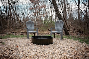 Two Adirondack chairs, a fire pit, and a gravel ground surrounded by trees.