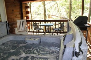 A hot tub with a wooden deck and chairs, surrounded by trees.