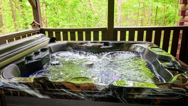 A hot tub with a waterfall feature, surrounded by a wooden deck and a forested backdrop. - Lazy Lane Cabins - Wren's Nest (Logan)