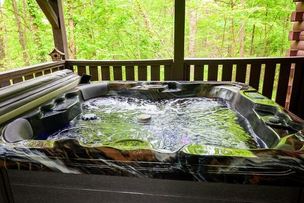 A hot tub with a waterfall feature, surrounded by a wooden deck and a forested backdrop.
