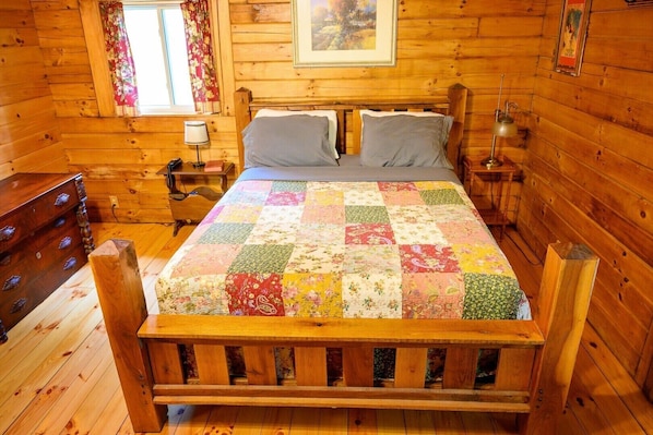 Un dormitorio de cabaña de madera con cama, cómoda, mesita de noche, lámpara y ventana con cortinas.