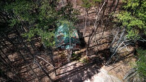 An aerial view of a cabin surrounded by dense forest, with a visible deck area and a small structure.