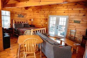 A cozy log cabin room with a bed, a dining table with chairs, a sofa, and a small table with a lamp.