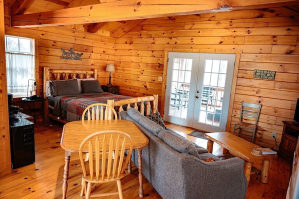 A cozy log cabin room with a bed, a dining table with chairs, a sofa, and a small table with a lamp.