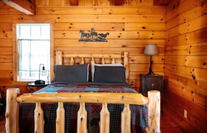 A wooden cabin bedroom with a bed, a nightstand, a lamp, and a window.