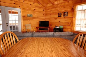 A wooden cabin interior with a dining table, chairs, a television, and a sofa.