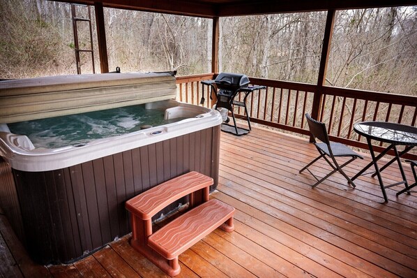 A hot tub on a wooden deck with a table and chairs, surrounded by trees.