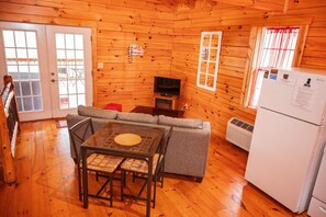 A cozy cabin interior with a sofa, a table with chairs, a television, and a refrigerator.