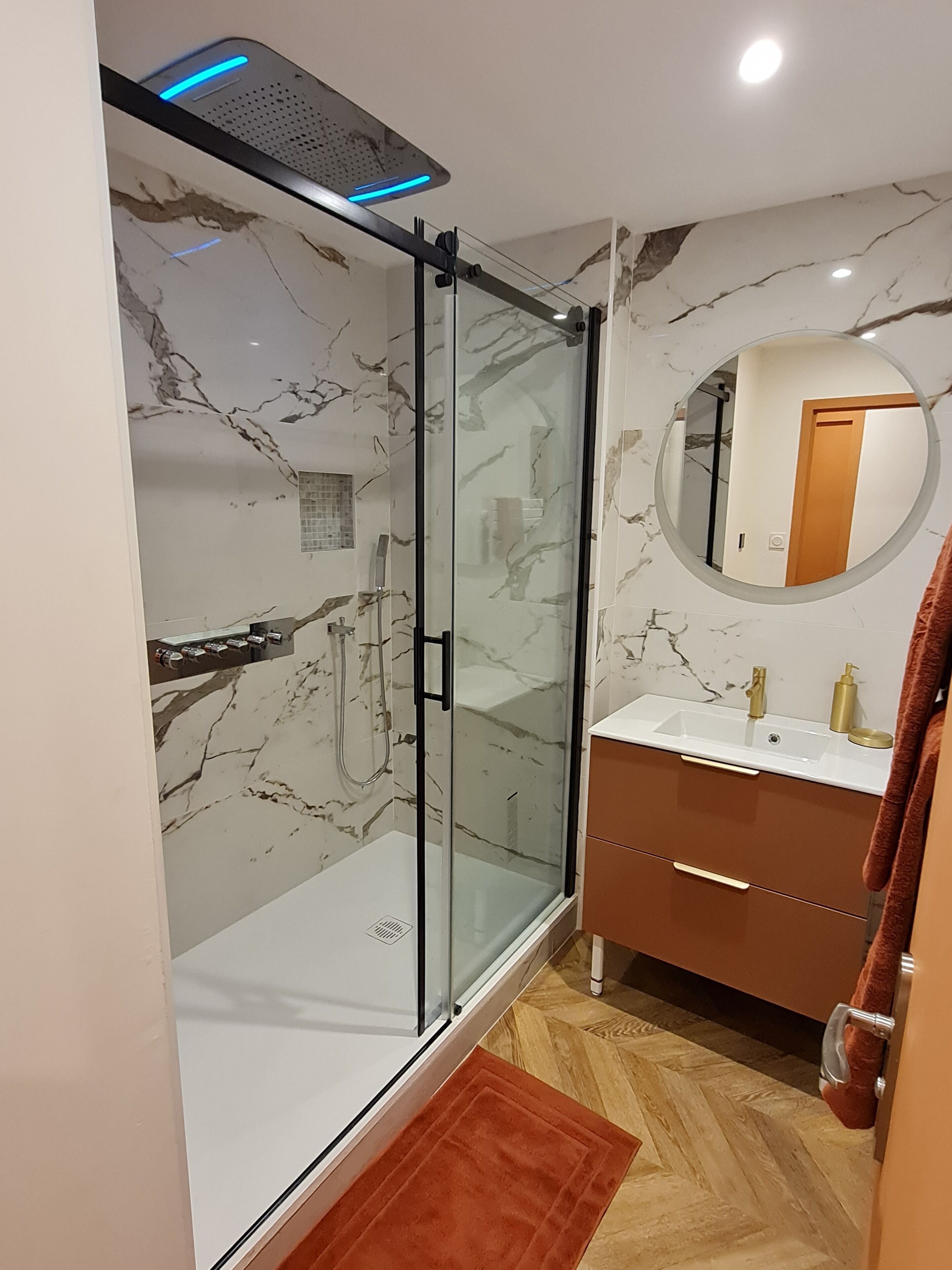 Ein modernes Badezimmer mit einer gläsernen Duschkabine, Marmorwänden, einem runden Spiegel und Holzboden.