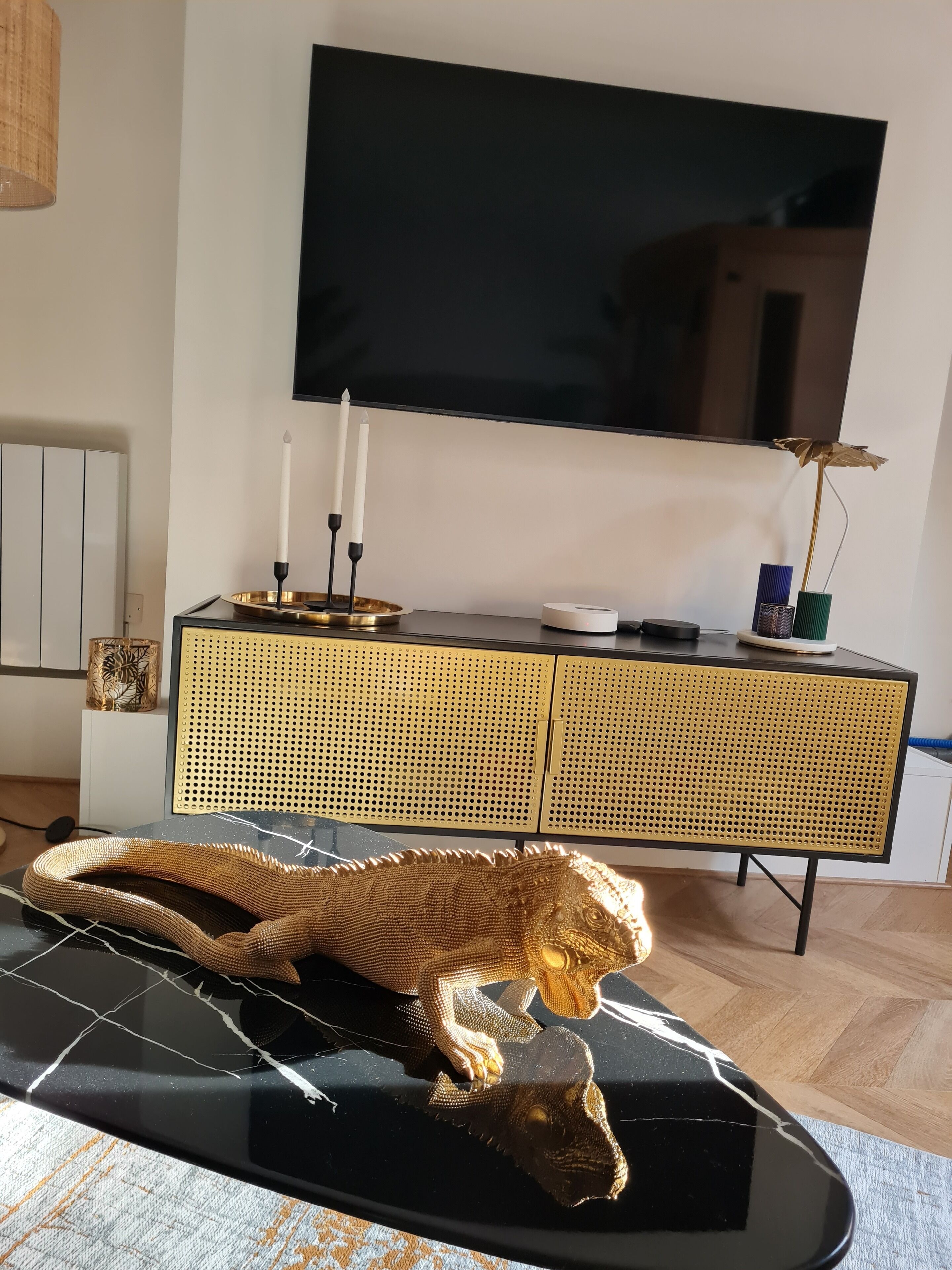 A room with a black leather couch, a television mounted on the wall, a woven cabinet, and a decorative plant.