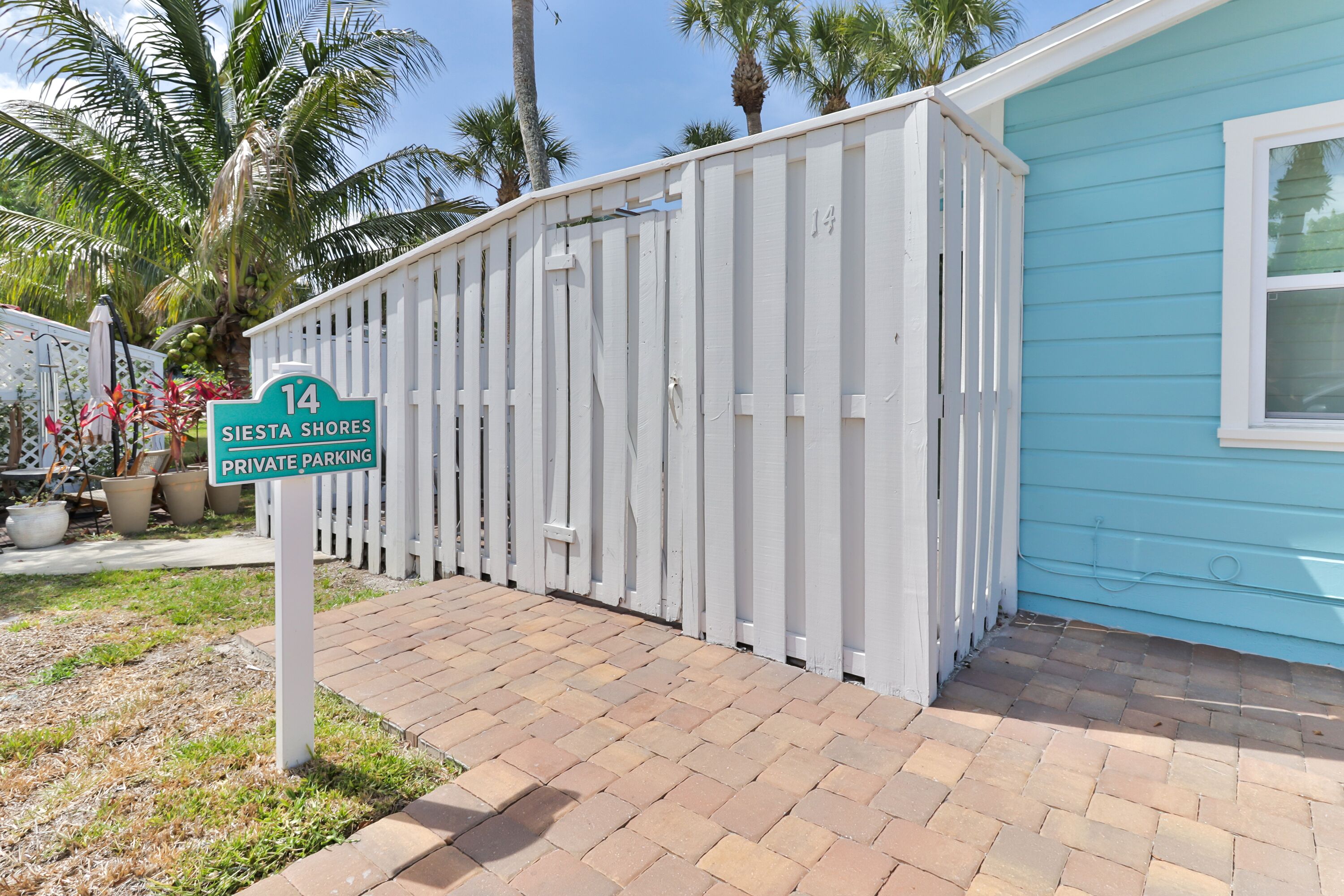 Un espace de stationnement privé délimité par une clôture blanche, avec un panneau indiquant « Stationnement privé – 14 Siesta Shores », et un bâtiment bleu en arrière-plan.