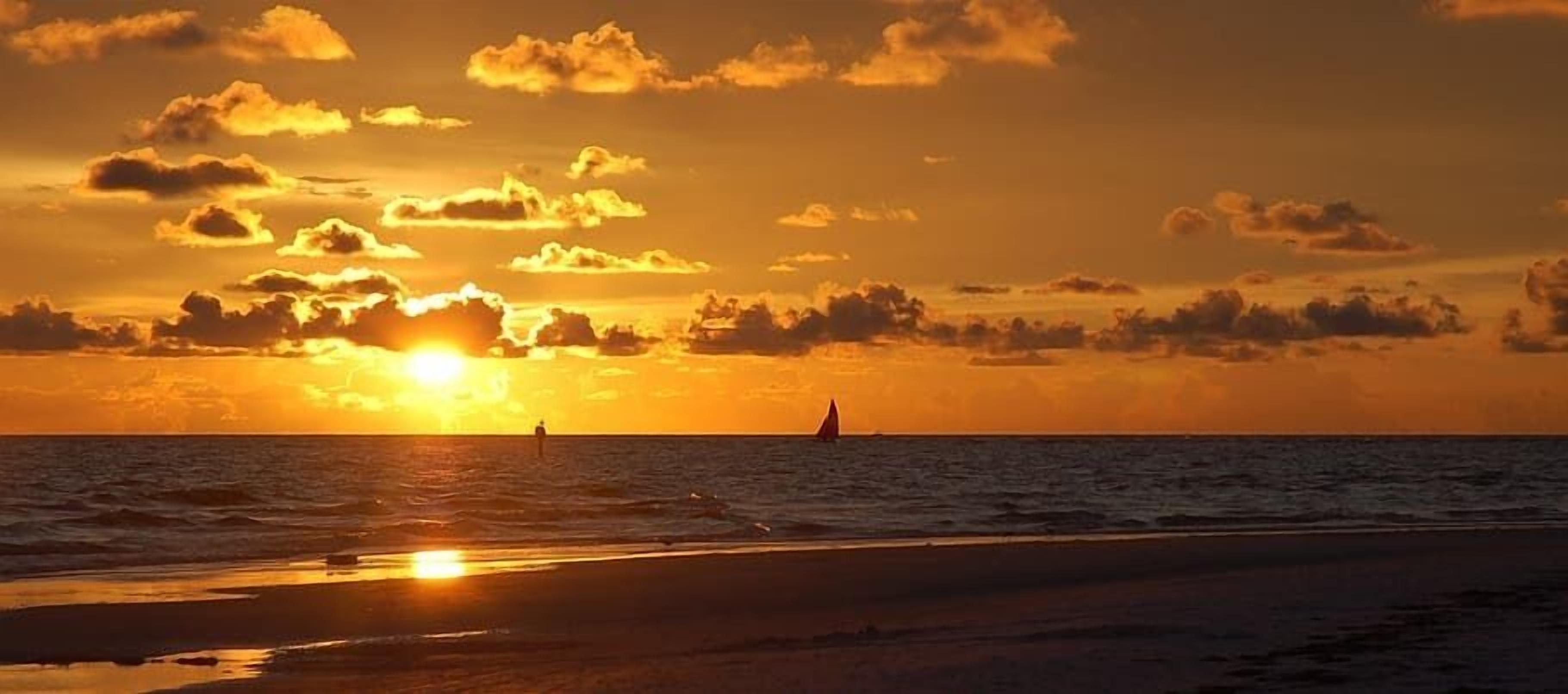 Un sol ponent-se sobre l'oceà amb un veler i una persona de peu a la platja.