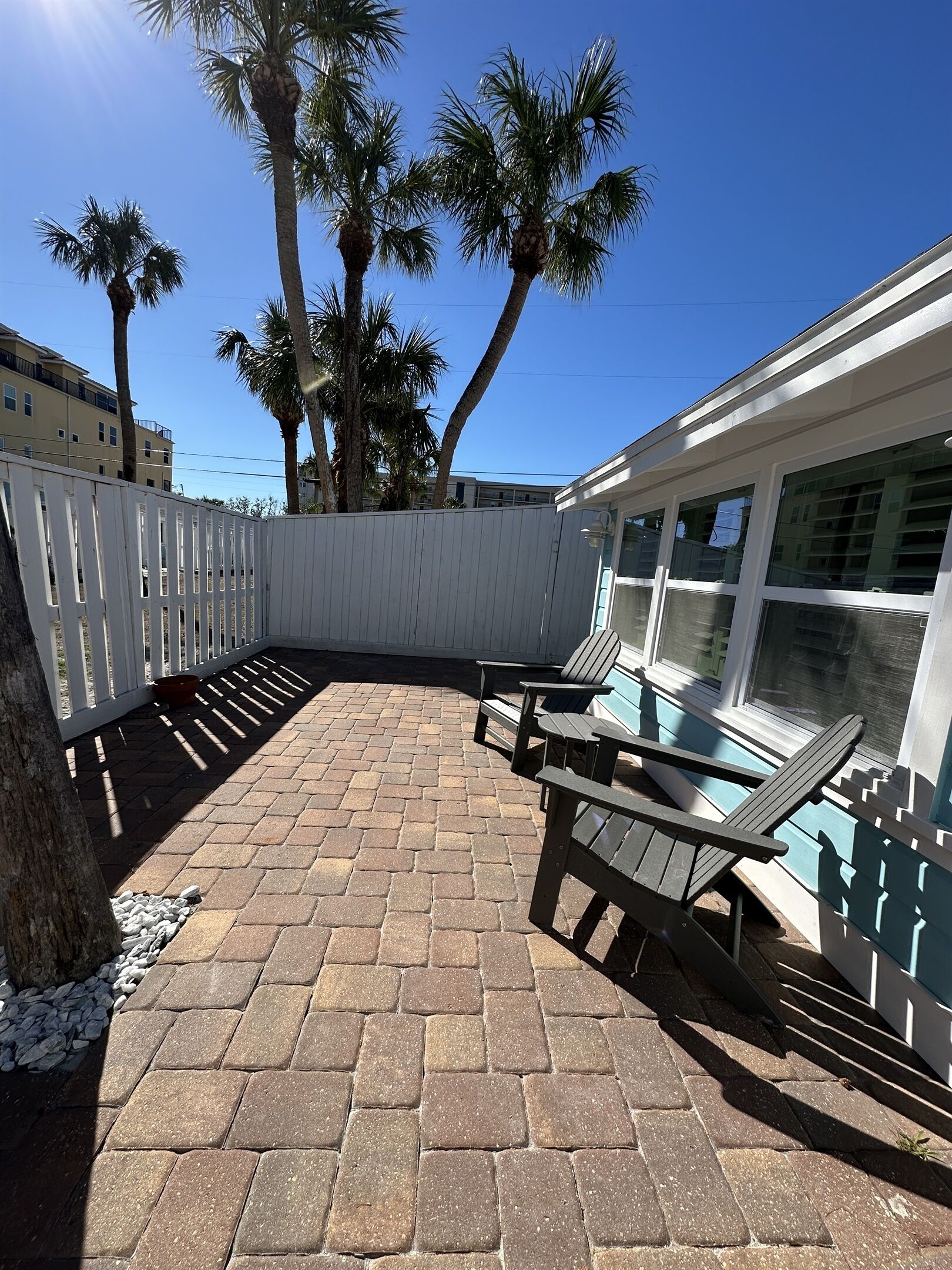 A patio area with a brick floor, wooden deck chairs, and a poolside area with a white fence and palm trees.