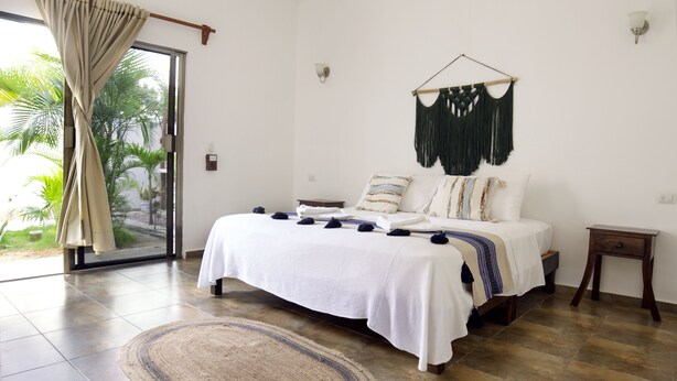 Un dormitorio con una cama grande, un adorno de macramé colgado en la pared, una mesita de noche pequeña y vistas a través de una puerta corrediza de vidrio.