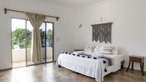 Un dormitorio con cama, mesita de noche, un adorno de macramé en la pared y una puerta corrediza de vidrio que da a un espacio exterior.