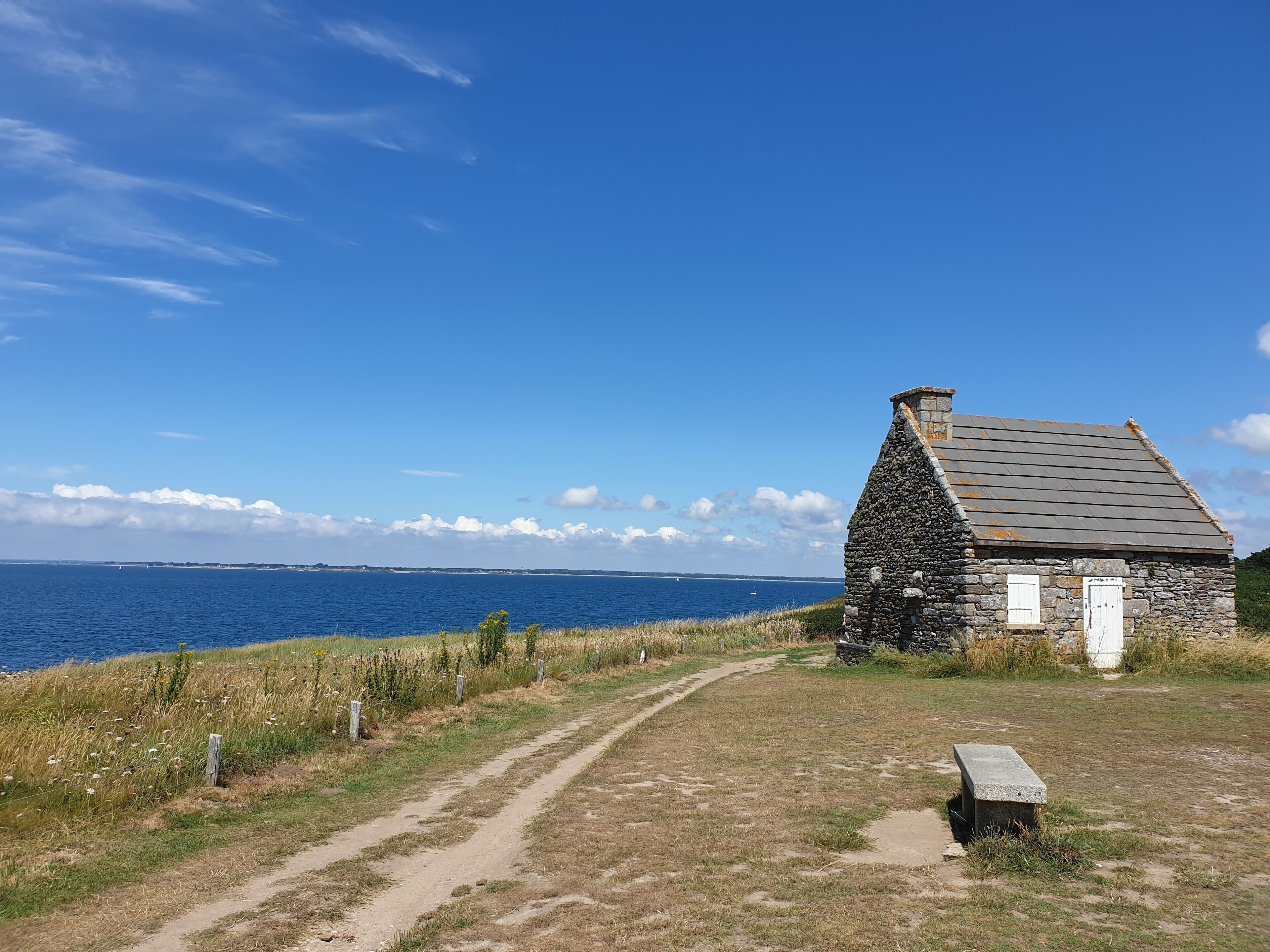 A stone cottage with a path leading to the sea, a bench, and a clear view of the ocean.