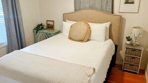 Cama bem arrumada com colcha branca, almofada de tecido trançado e cabeceira de madeira.