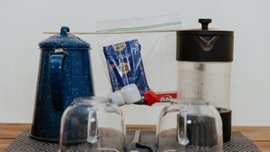 Eine Kaffeemaschine, ein French Press Kaffeefilter, ein Honigportion und eine Kaffeepackung auf einem Tisch.
