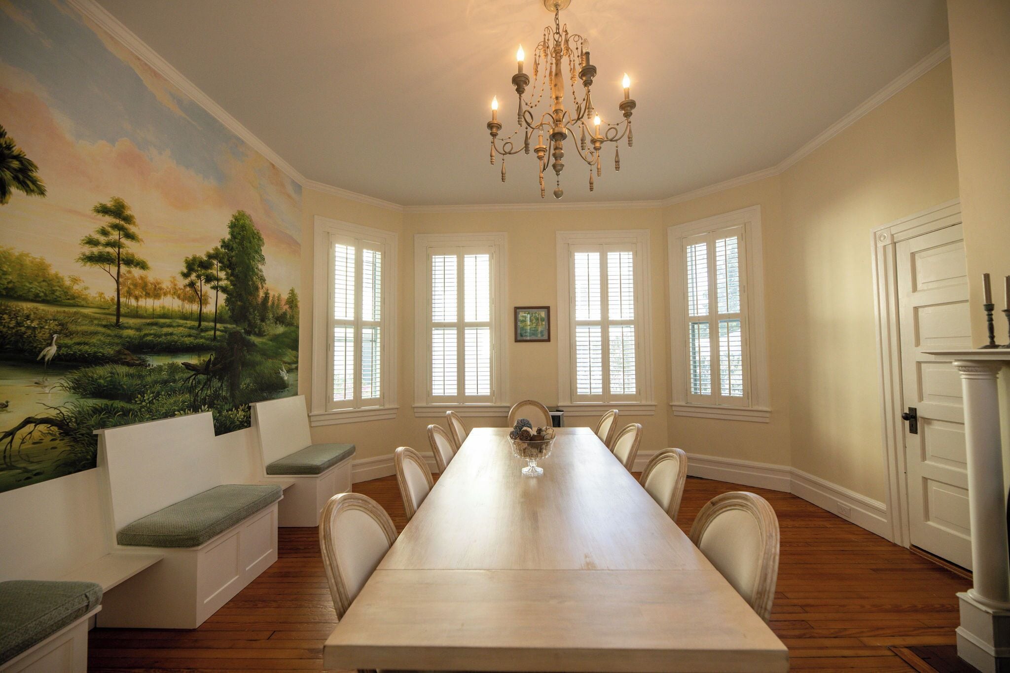 A dining room with a long table, white chairs, a chandelier, and a mural of a landscape.