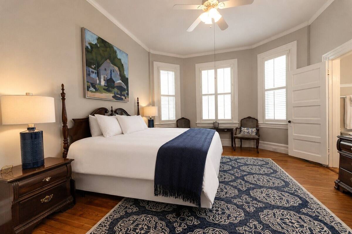 A bedroom with a large bed, a nightstand with a lamp, a dresser, a chair, and a painting on the wall.