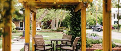 A covered patio with wicker chairs and a table, surrounded by greenery and a view of a street.