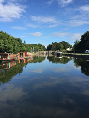A calm river with a wooden boat docked on the left, a stone bridge in the middle, and trees lining the riverbanks.