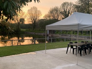 A tented outdoor area with a table and chairs, a pond, and trees in the background.