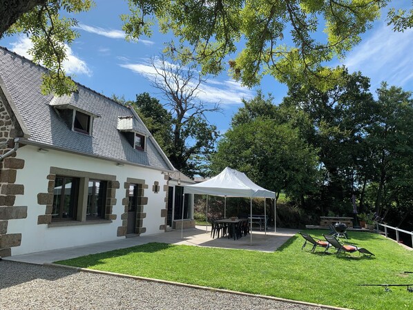 A house with stone walls and a white facade, a covered patio with tables and chairs, and a lawn with a grill and lounge chairs.