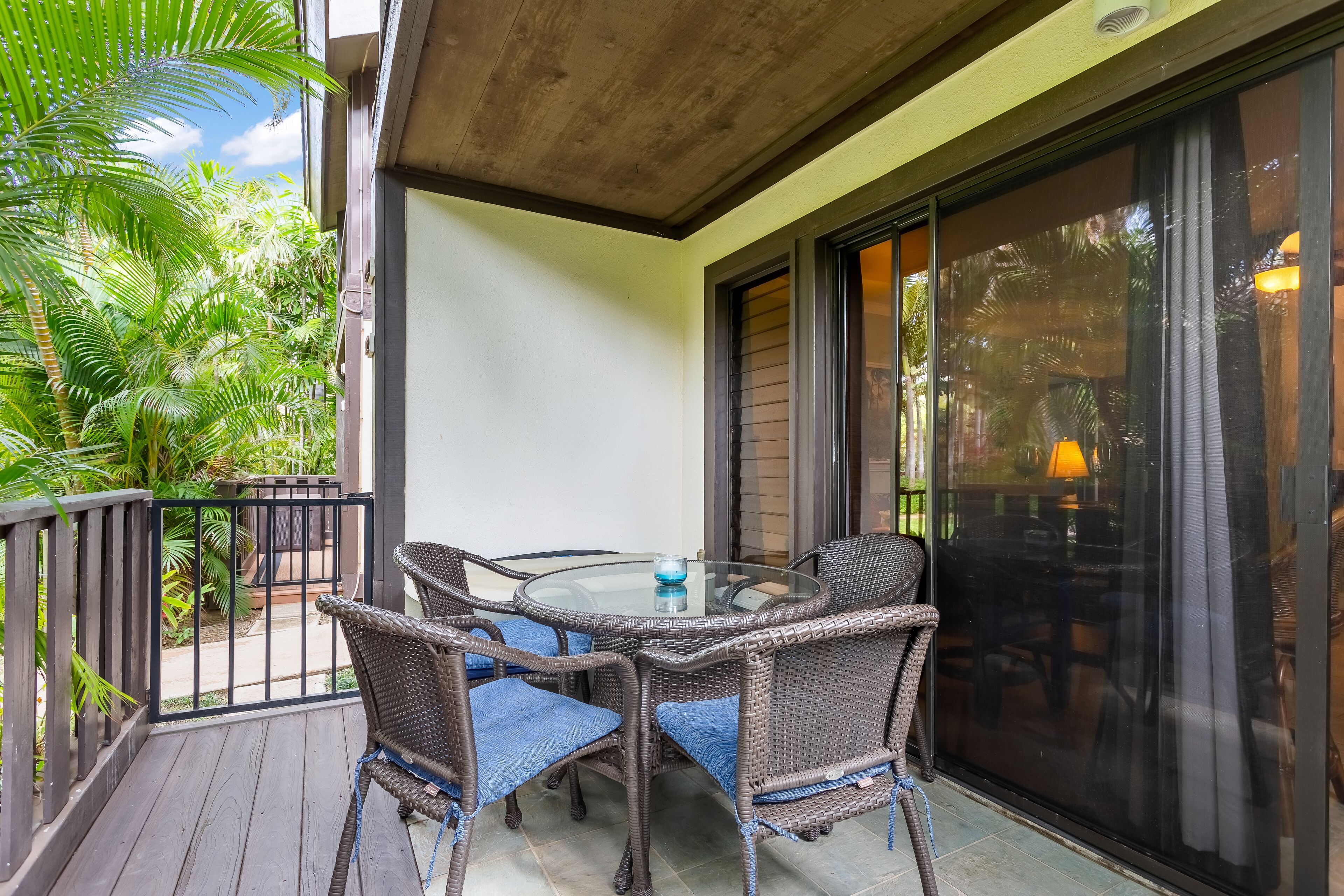 A balcony with wicker furniture and a glass table, surrounded by greenery.