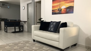 Un salon avec un canapé blanc, un tapis à motifs, un tableau représentant un coucher de soleil et un coin repas avec une table et des chaises.
