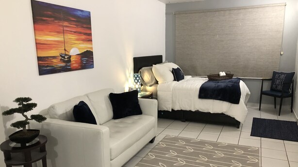 Une chambre moderne comprenant un lit, un canapé, un fauteuil, une petite table sur laquelle est posé un bonsaï, et un tableau représentant un coucher de soleil.