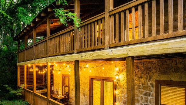 Une cabane en bois à deux étages, avec des murs en pierre, dotée d’un balcon décoré de guirlandes lumineuses, de meubles en bois et d’une cheminée en pierre.