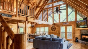 Un intérieur de cabine spacieux, avec un plafond haut, des poutres en bois, un coin repas, une cheminée et de grandes fenêtres.