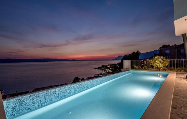 Relax by the serene pool overlooking a vast body of water and majestic mountains.