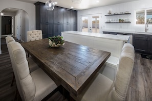 Une cuisine moderne avec des meubles de couleur sombre, un plan de travail blanc et une table à manger en bois, dotée de chaises rembourrées.