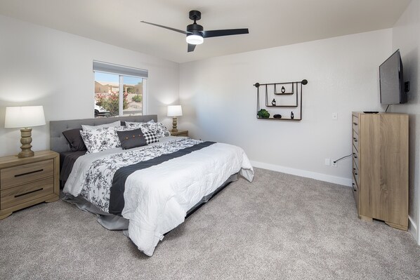 A bedroom with a bed, bedside tables, a ceiling fan, a TV, and a window with a view of buildings.