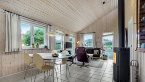 Een ruime kamer met houten plafond en wanden, een eettafel met stoelen, een bank, een open haard en grote ramen met gordijnen.