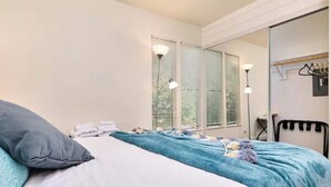 Un dormitorio con cama, ventana con persianas, lámpara de noche y espejo con perchero.