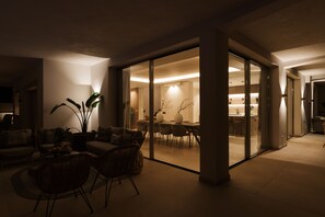 Una sala de estar moderna con comedor, que presenta un separador de vidrio, mobiliario de mimbre e iluminación ambiental.