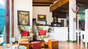 A cozy living area with floral-patterned armchairs, a wooden coffee table, and a red ottoman. There are framed artworks on the walls and a kitchen area in the background. - The Blue Suites & Spa (La Fortuna)