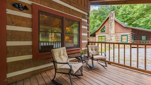 A wooden deck with rocking chairs and a table, overlooking a cabin with a stone chimney and green shutters. - Cozy2 BD Easily Accessible Cabin! Quiet community short drive to all attractions (Pigeon Forge)