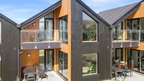 A modern two-story house with large windows, a balcony, and a patio area with outdoor furniture.