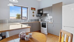 A modern kitchen with a wooden dining table, a refrigerator, a stove, and a window with a view of palm trees. - Ekhaya - New Plymouth Holiday Home (New Plymouth)