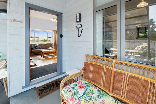 Un espacio de sala acogedor con un sofá de mimbre, una mesita y un banco. Una puerta da a un patio exterior con un sofá y una mesa.