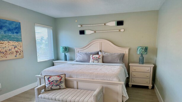 A bedroom with a bed, nightstands, a bench, a painting, and a window with blinds.