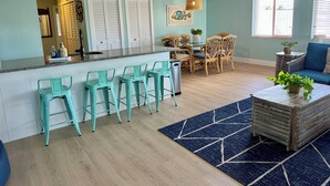 Eine moderne Küche mit türkisfarbenen Barhocker, ein Essbereich mit Korbstühlen und ein Wohnzimmer mit blauem Sofa.